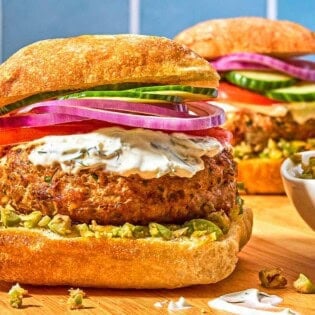 Two turkey burgers on ciabatta rolls dressed with zeytoon parvardeh, tzatziki, tomatoes, red onions, and cucumbers next to a bowl of the zeytoon parvardeh.