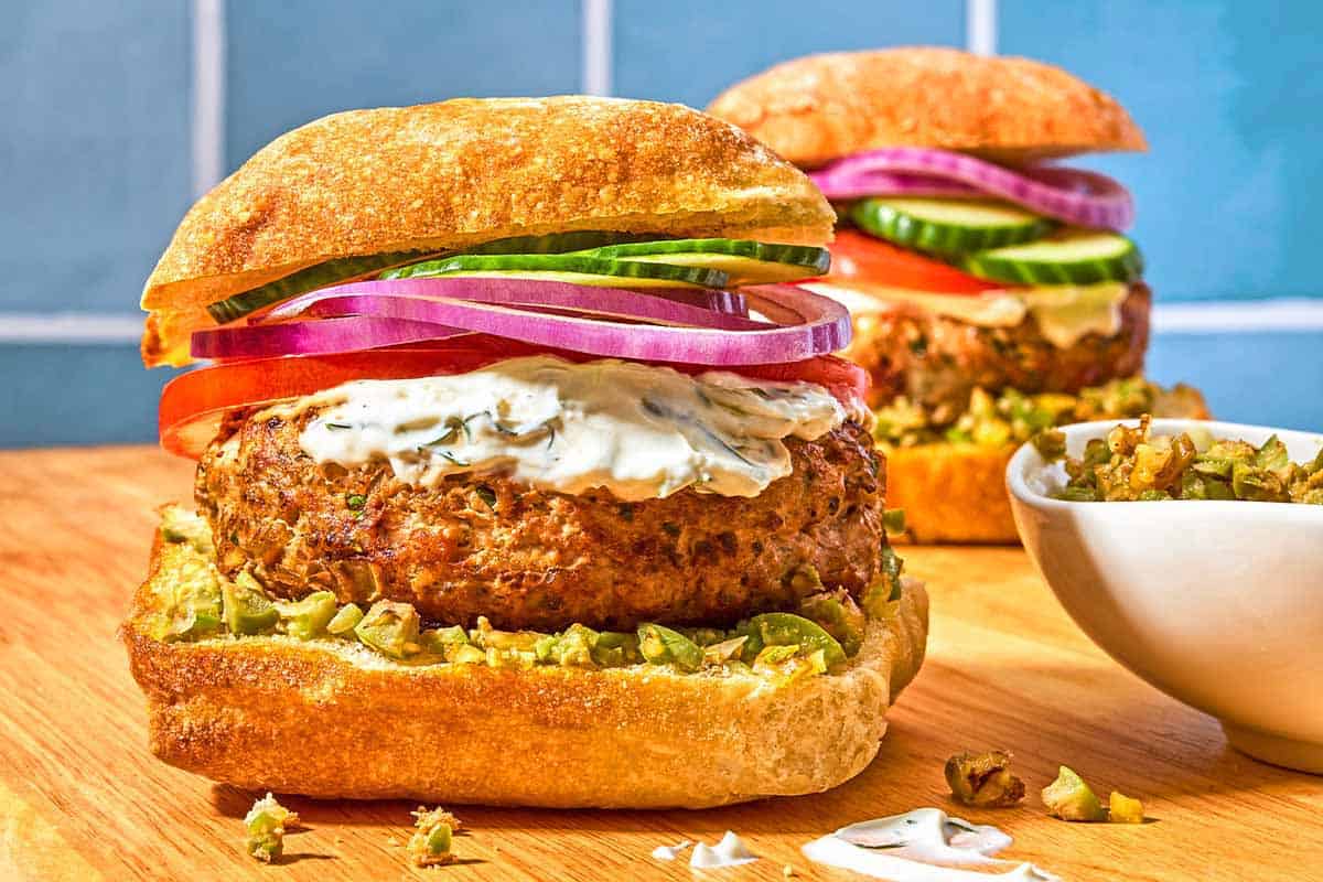 Two turkey burgers on ciabatta rolls dressed with zeytoon parvardeh, tzatziki, tomatoes, red onions, and cucumbers next to a bowl of the zeytoon parvardeh.