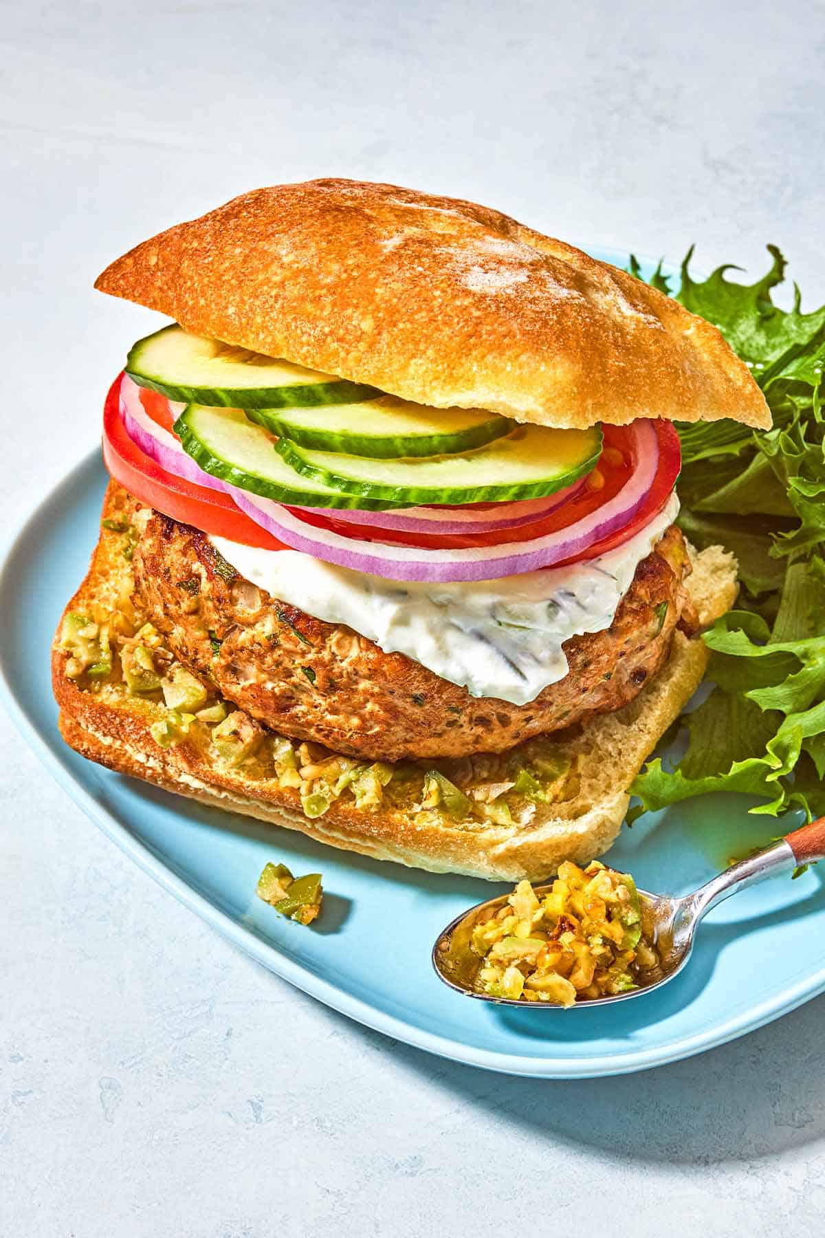 A turkey burger on a ciabatta roll dressed with zeytoon parvardeh, tzatziki, tomatoes, red onions, and cucumbers on a plate with lettuce and a spoonful of zeytoon parvardeh.