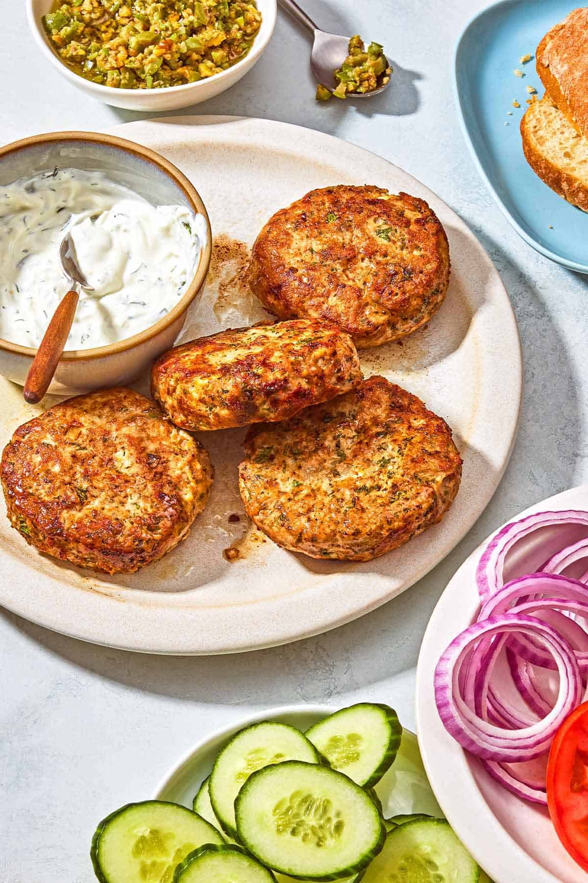 A plate with 4 plain turkey burger patties and a bowl of tzatziki with a spoon. Next to this is a bowl of zeytoon parvardeh, a spoon, a plate with a ciabatta roll, and plates with slices of red onion, tomato and cucumber.