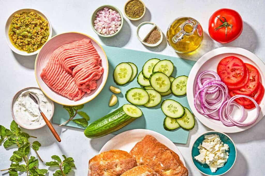 Ingredients for turkey burgers including ground turkey, a shallot, mint leaves, feta, garlic, oregano, salt, black pepper, olive oil, rolls, tomatoes, cucumber, red onion tzatziki, zeytoon parvardeh.