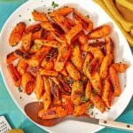 The roasted carrot recipe on a platter with a serving spoon surrounded by a bowl of chopped parsley, a lemon, a bowl of sumac, and a cloth napkin.