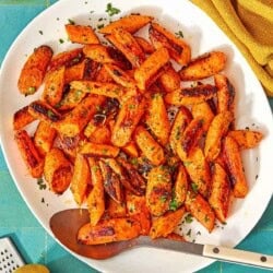 The roasted carrot recipe on a platter with a serving spoon surrounded by a bowl of chopped parsley, a lemon, a bowl of sumac, and a cloth napkin.