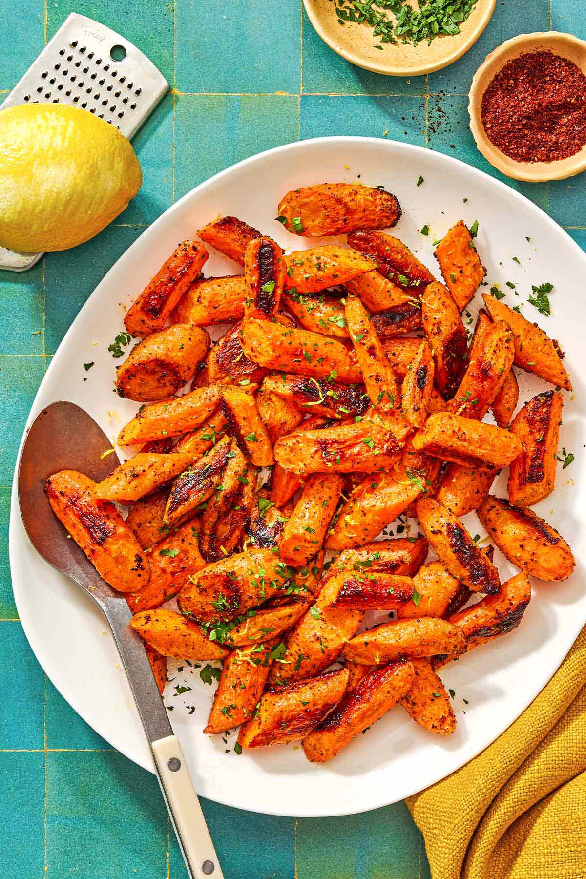 The roasted carrot recipe on a platter with a serving spoon surrounded by a bowl of chopped parsley, a lemon, a bowl of sumac, and a cloth napkin.
