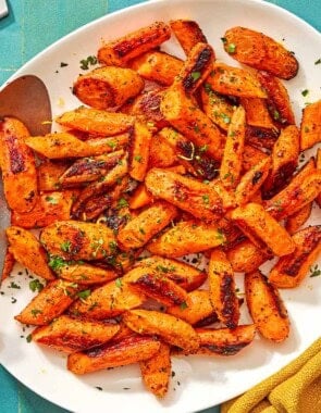 The roasted carrot recipe on a platter with a serving spoon surrounded by a bowl of chopped parsley, a lemon, a bowl of sumac, and a cloth napkin.