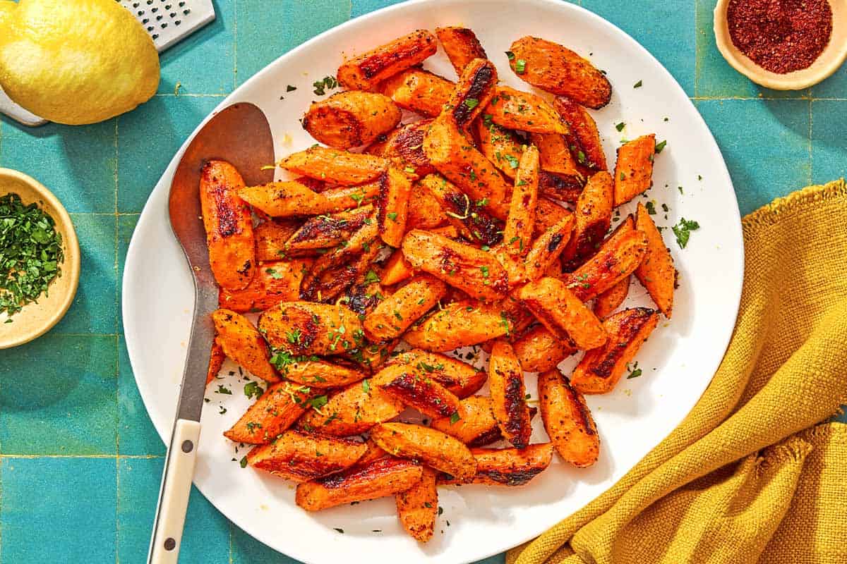 The roasted carrot recipe on a platter with a serving spoon surrounded by a bowl of chopped parsley, a lemon, a bowl of sumac, and a cloth napkin.