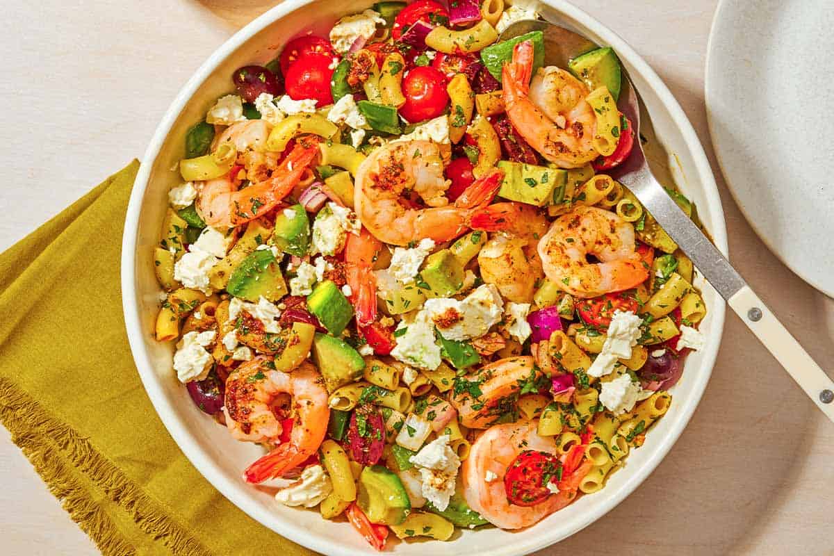 A bowl of shrimp pasta salad with a serving spoon next to a cloth napkin and a plate.