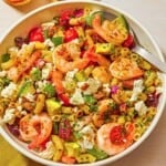 The shrimp pasta salad in a bowl with a spoon next to a cloth napkin, a bowl of feta and a glass of sparkling water.