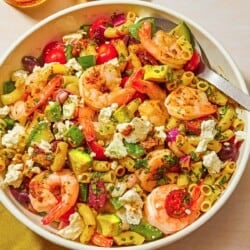 The shrimp pasta salad in a bowl with a spoon next to a cloth napkin, a bowl of feta and a glass of sparkling water.