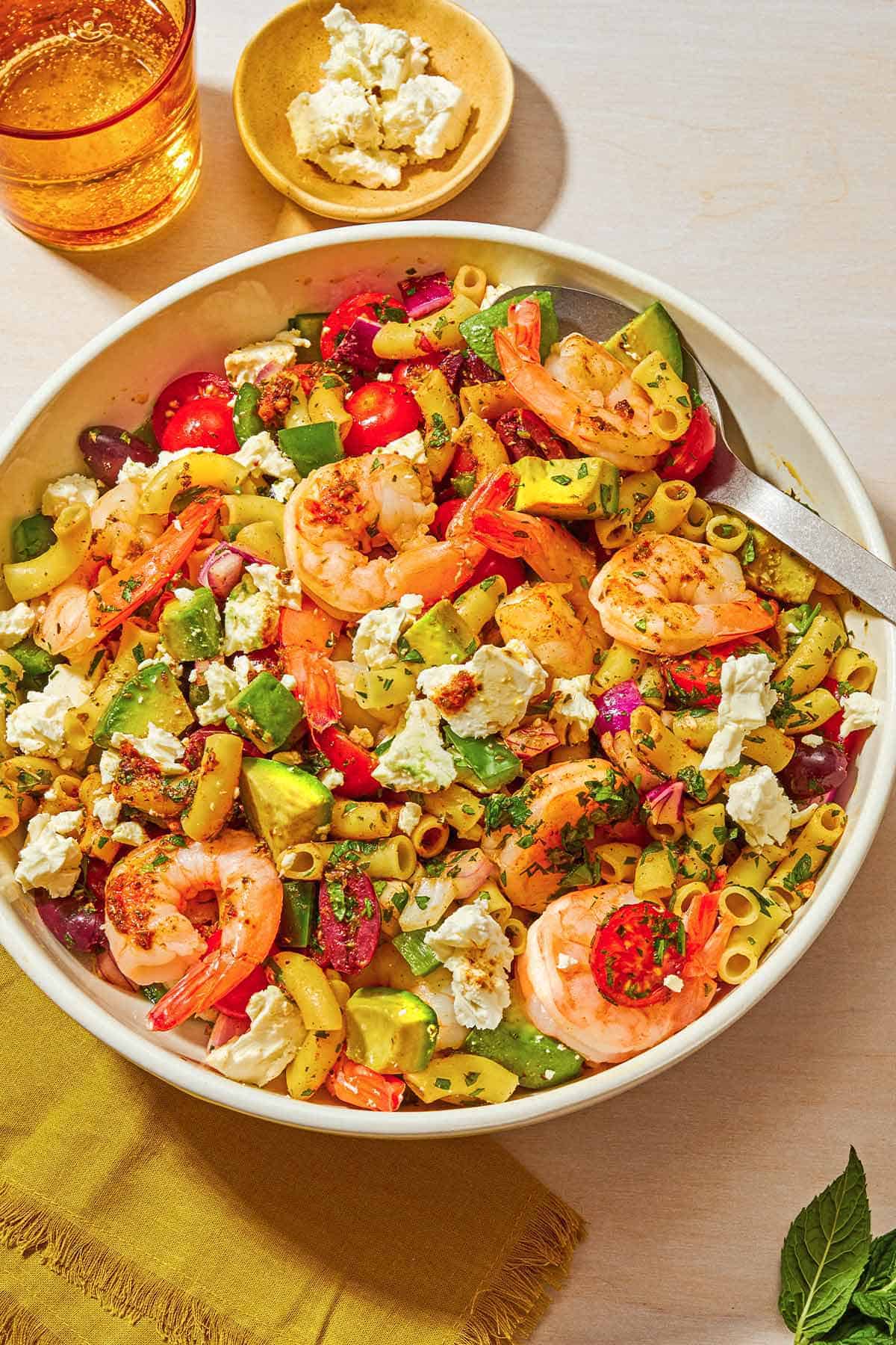 The shrimp pasta salad in a bowl with a spoon next to a cloth napkin, a bowl of feta and a glass of sparkling water.