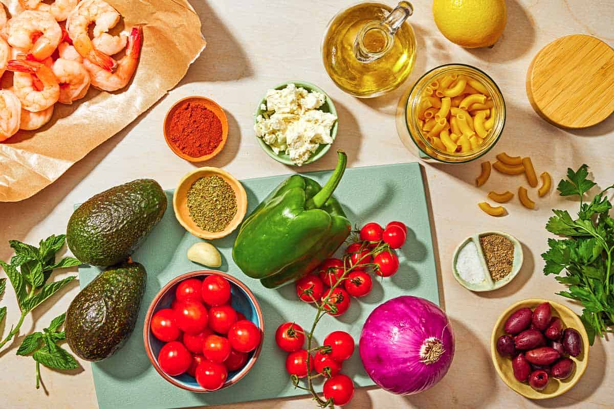 The ingredients for the shrimp pasta salad including shrimp, elbow macaroni, green bell pepper, cherry tomatoes, red onion, kalamata olives, parsley, mint, avocados, feta, lemon, olive oil, garlic, oregano, sweet paprika, salt and black pepper.