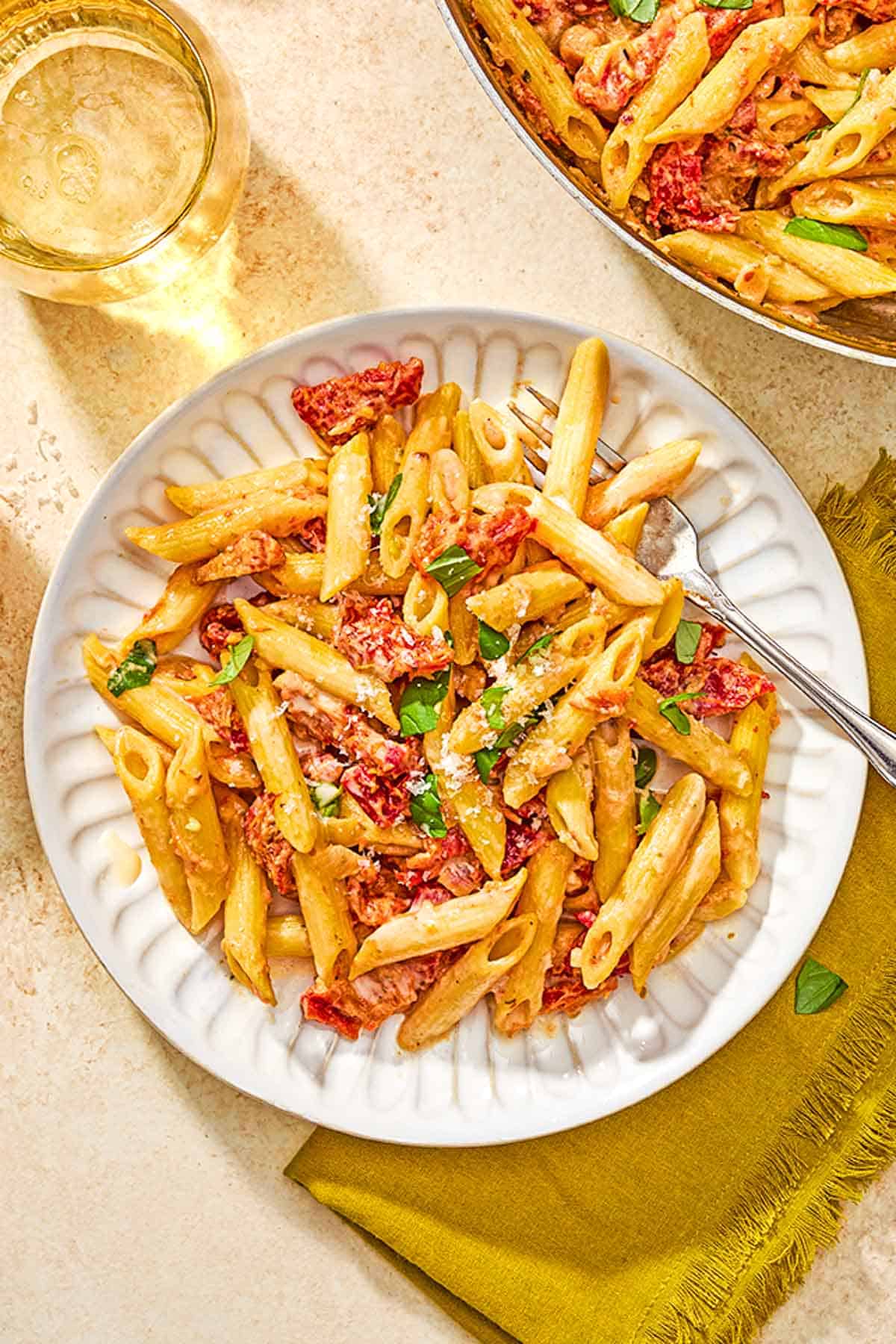 Sun dried tomato pasta on a plate with a fork next to a glass of white wine, a cloth napkin and the rest of the pasta in a skillet.