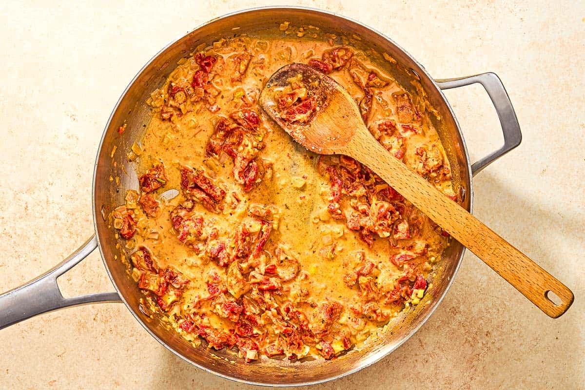 The shallots, garlic, sun dried tomatoes and milk cooking together in a skillet with a wooden spoon.