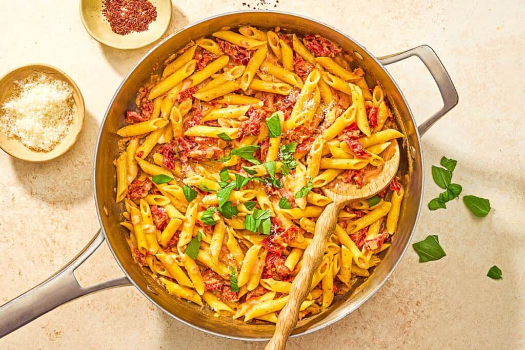 Sun dried tomato pasta in a skillet with a wooden spoon next to bowls of aleppo pepper and grated parmesan cheese.