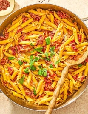 Sun dried tomato pasta in a skillet with a wooden spoon next to bowls of aleppo pepper and grated parmesan cheese.
