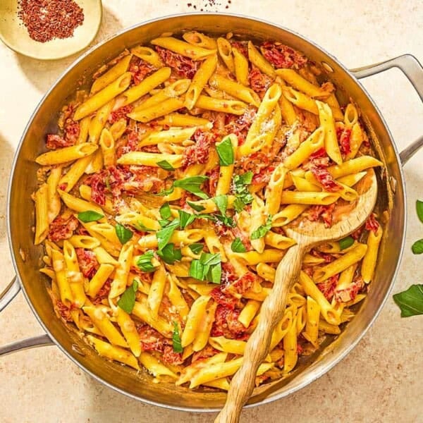 Sun dried tomato pasta in a skillet with a wooden spoon next to bowls of aleppo pepper and grated parmesan cheese.