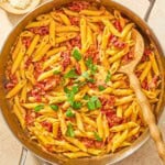 Sun dried tomato pasta in a skillet with a wooden spoon next to bowls of aleppo pepper and grated parmesan cheese.
