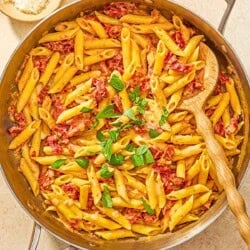 Sun dried tomato pasta in a skillet with a wooden spoon next to bowls of aleppo pepper and grated parmesan cheese.