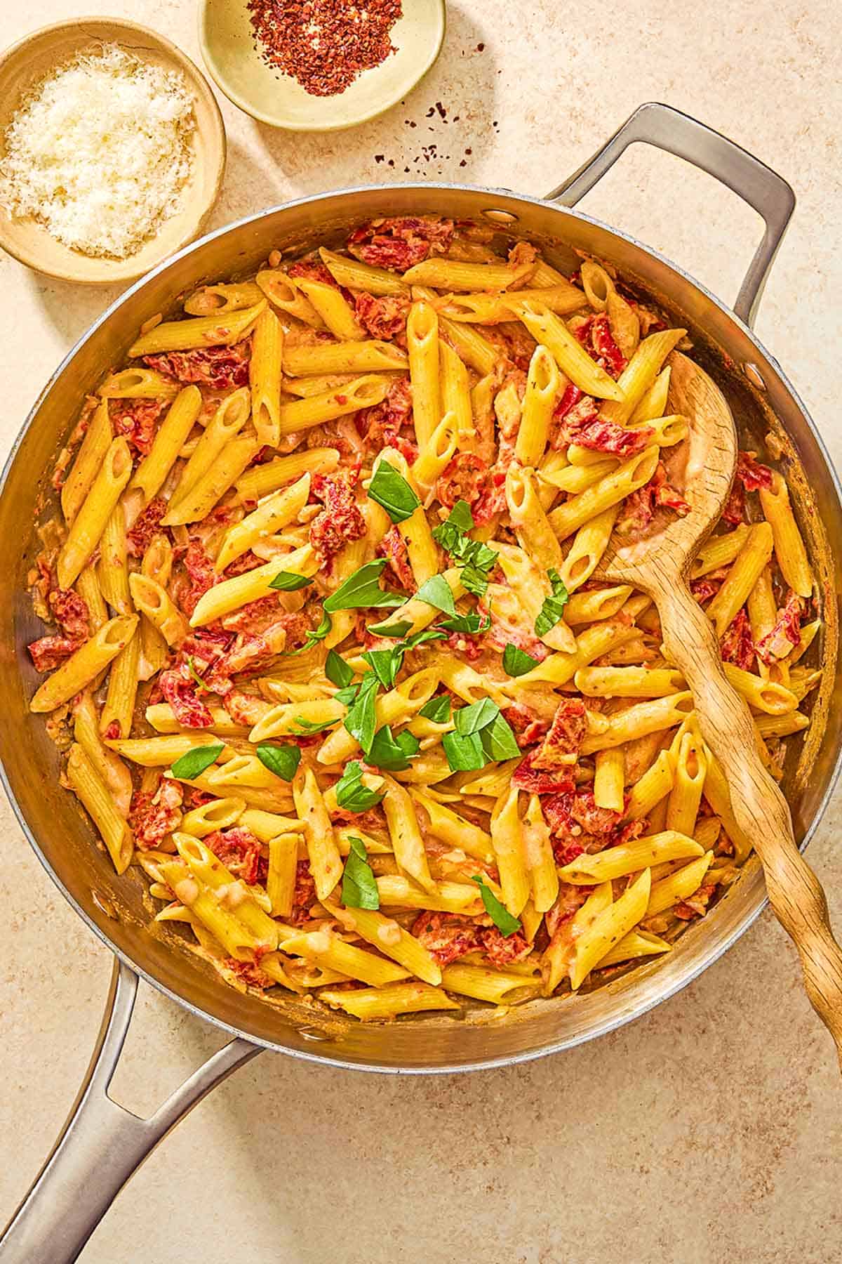 Sun dried tomato pasta in a skillet with a wooden spoon next to bowls of aleppo pepper and grated parmesan cheese.