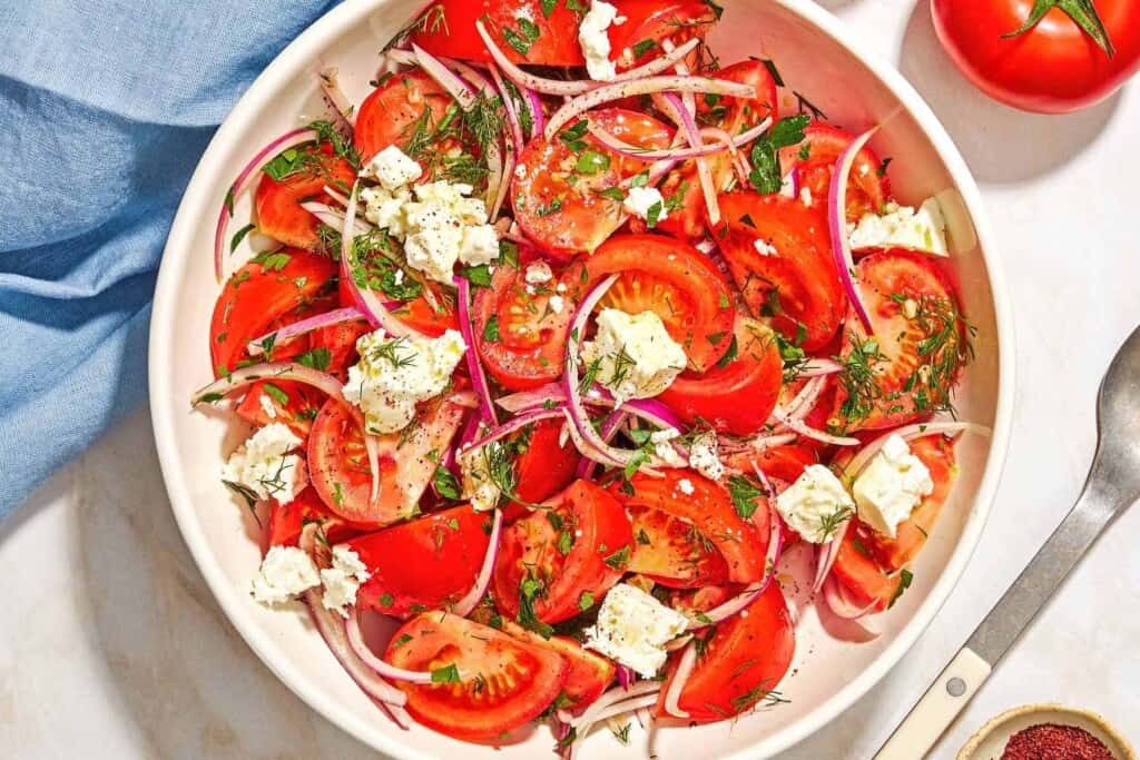 The tomato feta salad in a serving bowl next to a serving spoon, a bowl of sumac, a tomato, and a cloth napkin.