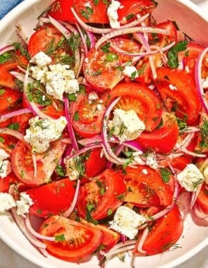 The tomato feta salad in a serving bowl next to a serving spoon, a bowl of sumac, a tomato, and a cloth napkin.