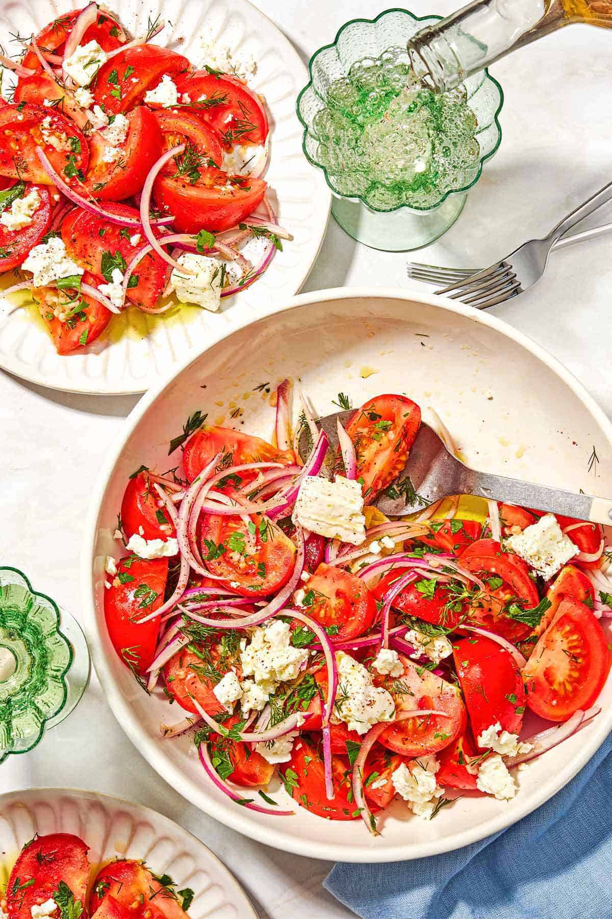 The tomato feta salad in a serving bowl with a serving spoon. Next to this is a cloth napkin, 2 glasses of white wine, two forks, and 2 servings of the salad on plates.