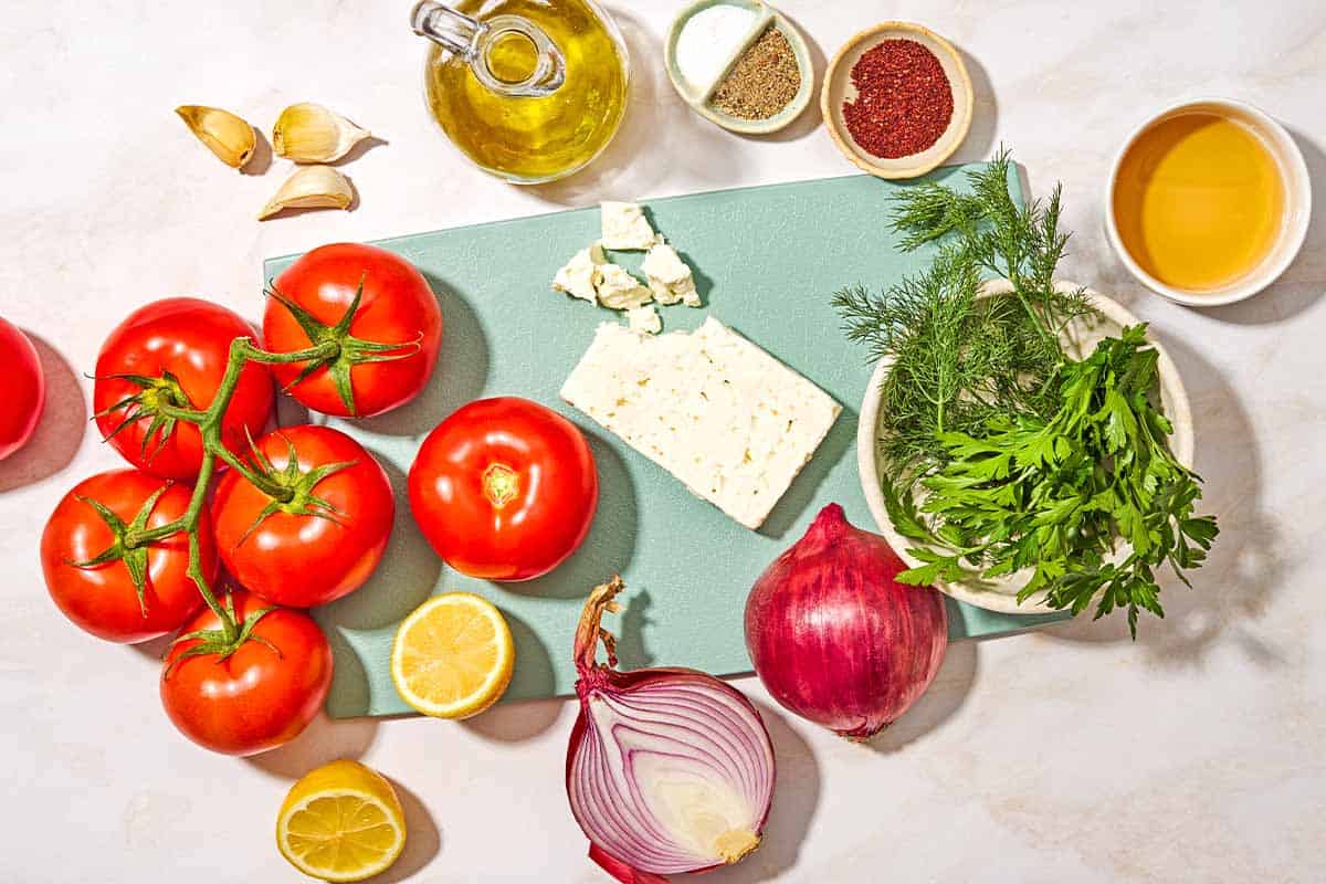 Ingredients for tomato feta salad including tomatoes, feta cheese, red onions, lemon, salt, black pepper, white wine vinegar, garlic, dill, parsley, and olive oil.