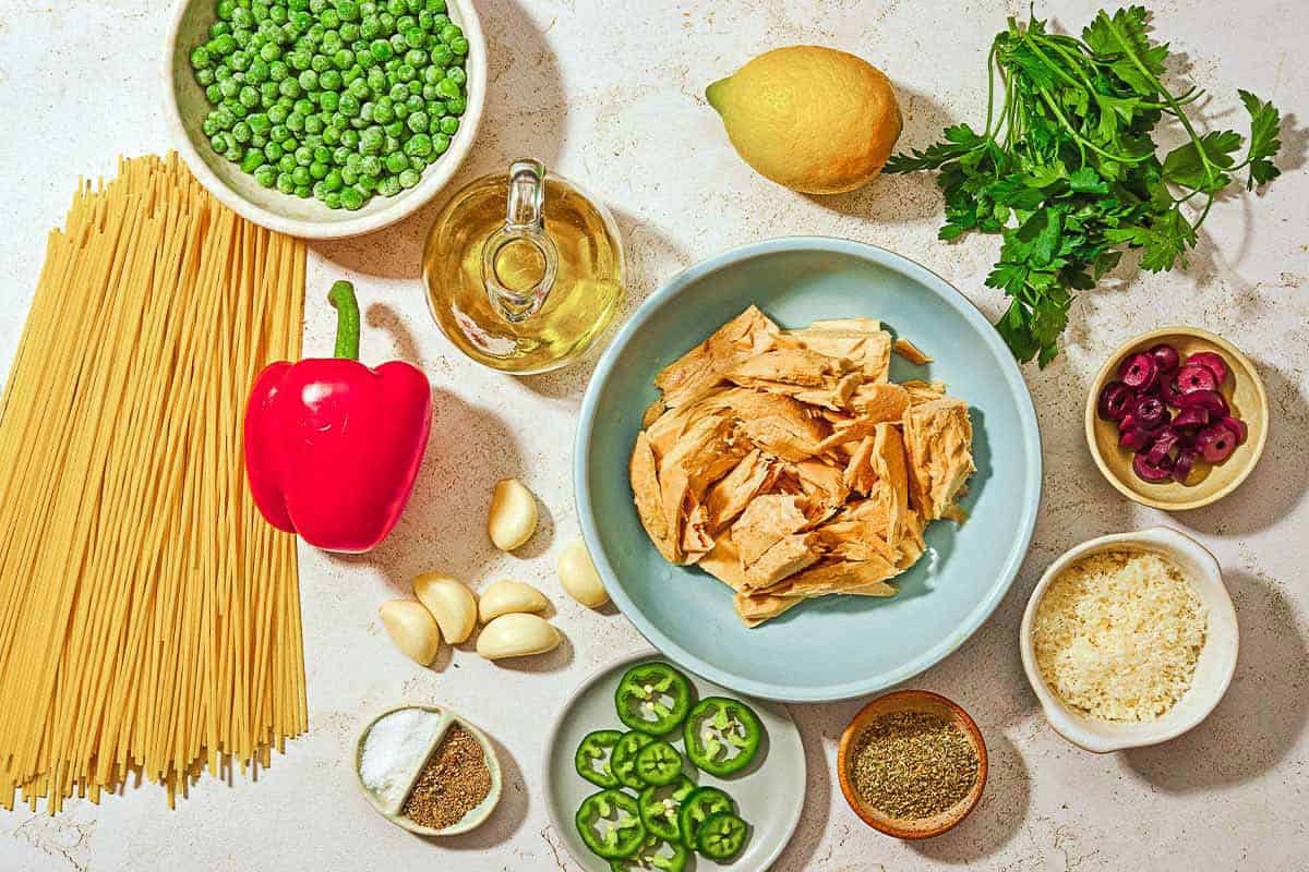 Ingredients for tuna pasta including spaghetti canned tuna, salt, frozen peas, olive oil, red bell pepper, garlic, lemon, parsley, dried oregano, black pepper, kalamata olives, jalapeno pepper, and grated Parmesan cheese.
