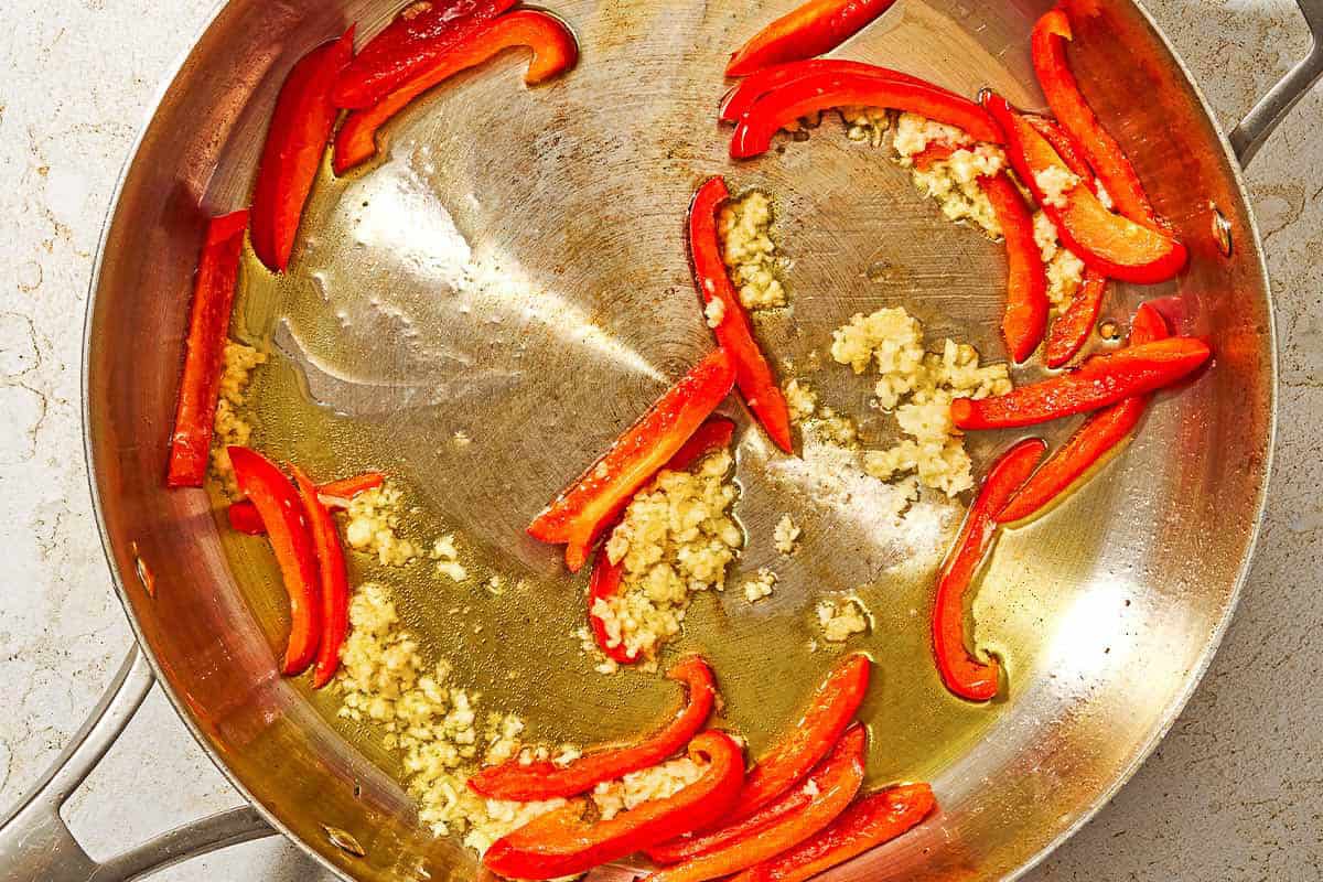 Red bell pepper strips and garlic being sauteed in olive oil in a deep skillet.