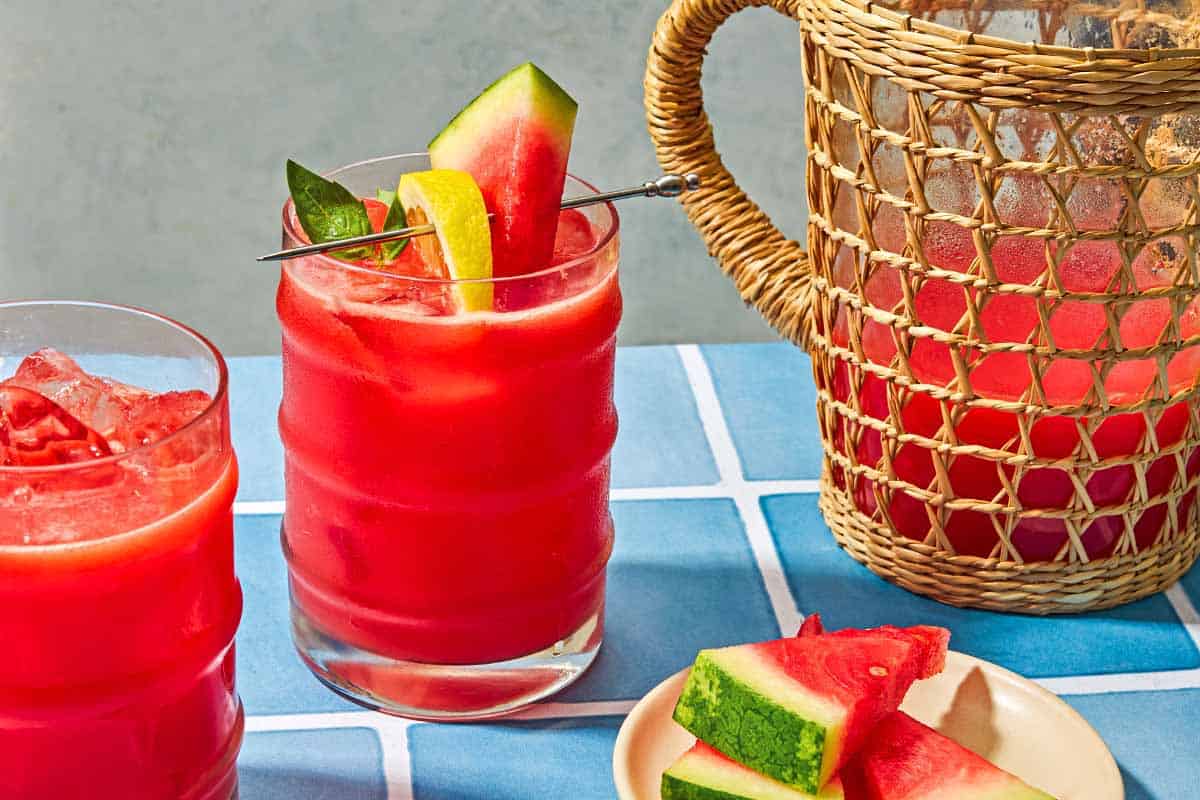 Two glasses of watermelon lemonade, one is garnished with basil and slices of watermelon and lemon. Next to these is a pitcher of the watermelon lemonade and a plate of sliced watermelon.