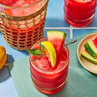 Two glasses of watermelon lemonade garnished with basil and slices of watermelon and lemon. Next to these is a pitcher of the watermelon lemonade and a plate of sliced watermelon.