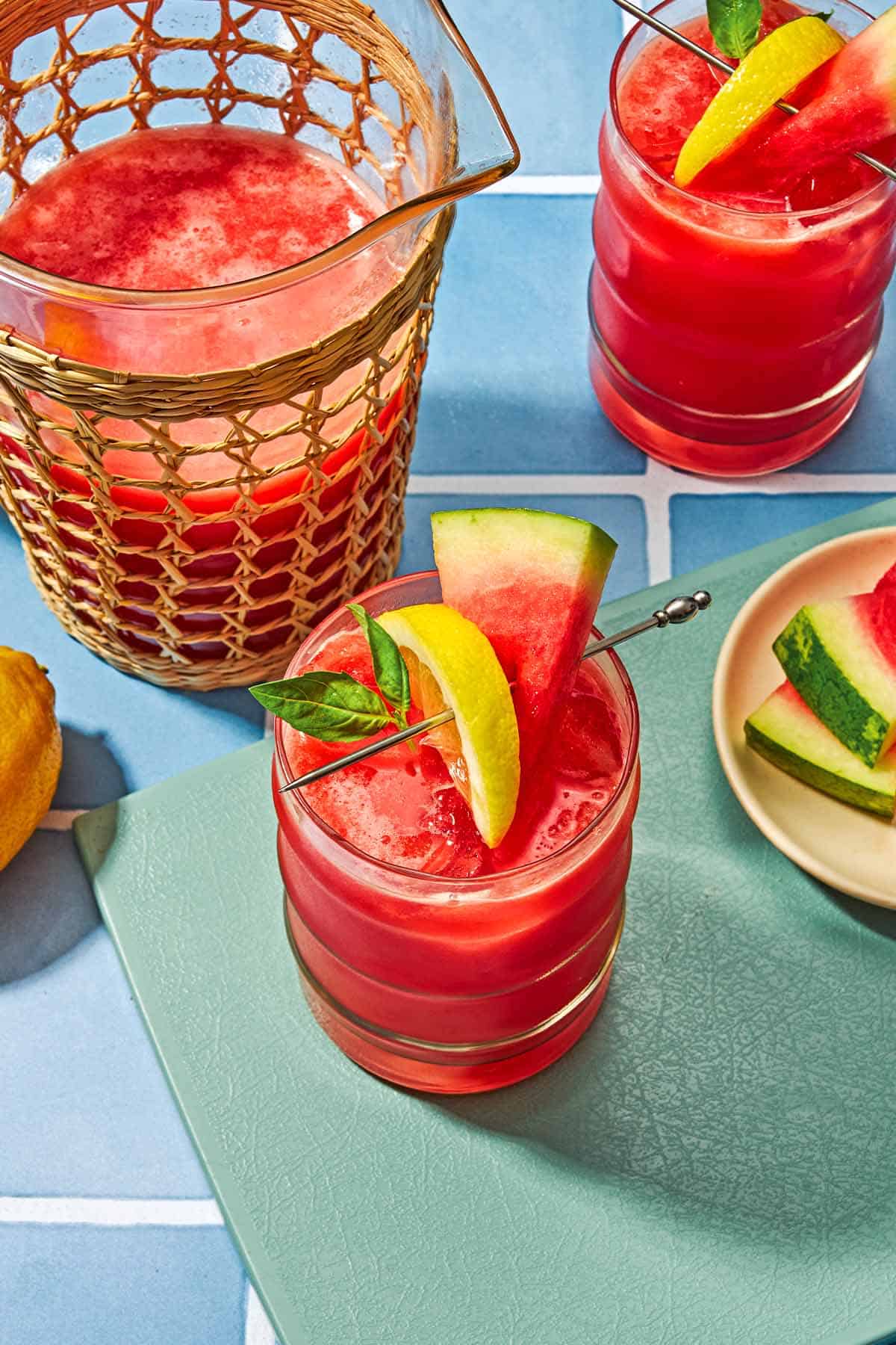 Two glasses of watermelon lemonade garnished with basil and slices of watermelon and lemon. Next to these is a pitcher of the watermelon lemonade and a plate of sliced watermelon.