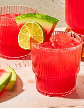 A glass of watermelon sangria garnished with a lime wheel and a watermelon slice next to another glass of the sangria, a pitcher of the sangria, a lime half and several slices of watermelon.