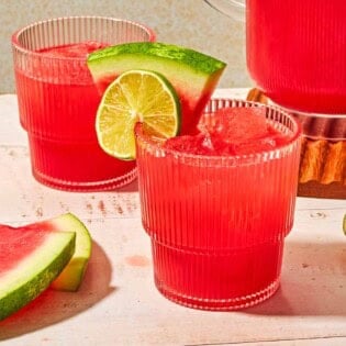 A glass of watermelon sangria garnished with a lime wheel and a watermelon slice next to another glass of the sangria, a pitcher of the sangria, a lime half and several slices of watermelon.