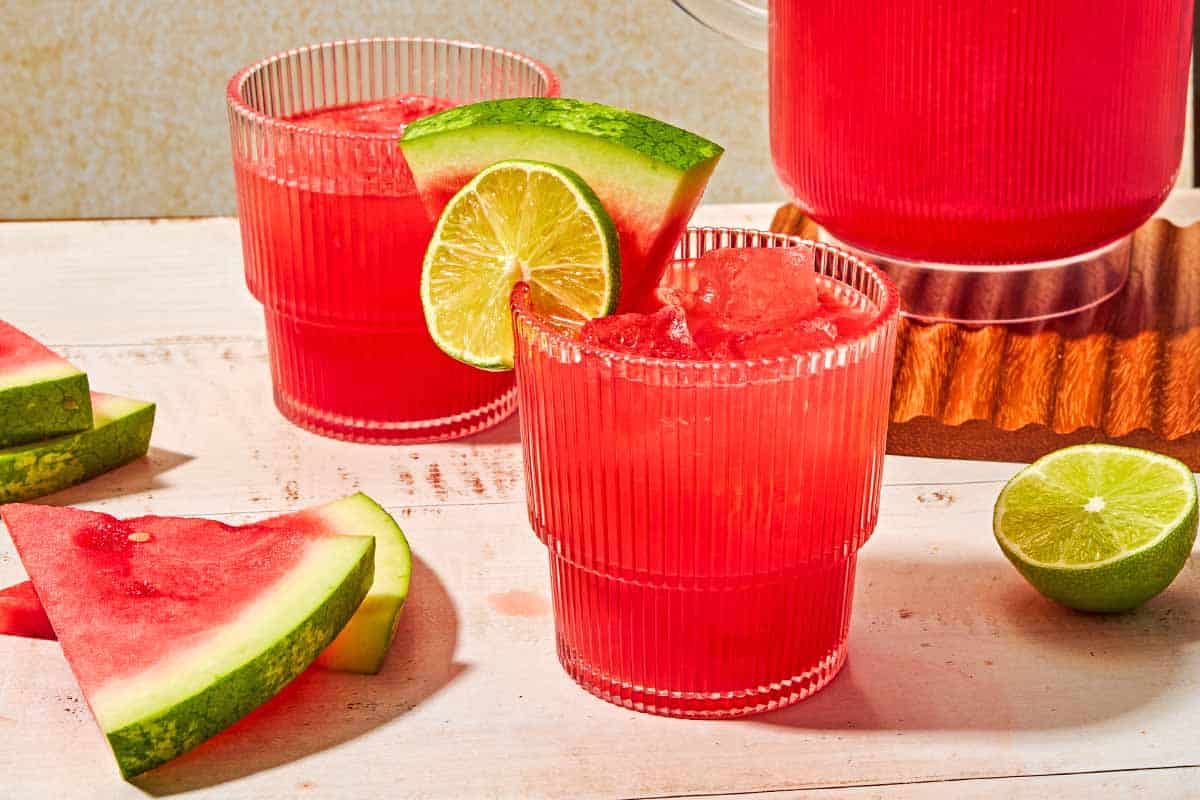 A glass of watermelon sangria garnished with a lime wheel and a watermelon slice next to another glass of the sangria, a pitcher of the sangria, a lime half and several slices of watermelon.