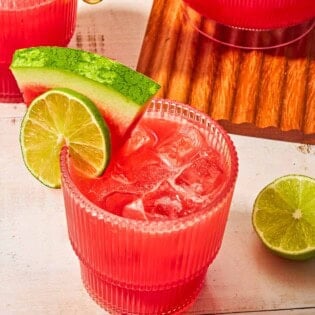 A glass of watermelon sangria garnished with a lime wheel and a watermelon slice next to another glass of the sangria, a pitcher of the sangria, and a lime half.