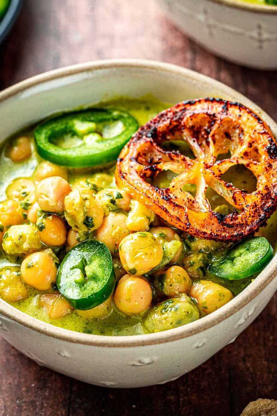 close up of two bowls of creamy pesto braised chickpeas topped with jalapeno and fried lemon slices, next to a plate of sliced jalapeno.
