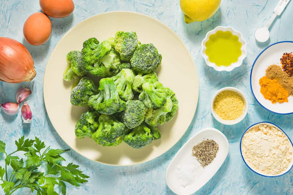 Ingredients for broccoli fritters including frozen broccoli, yellow onion, parsley, garlic, nutritional yeast, coriander, aleppo pepper, turmeric, baking powder, salt, black pepper, chickpea flour, eggs, olive oil, and lemon.