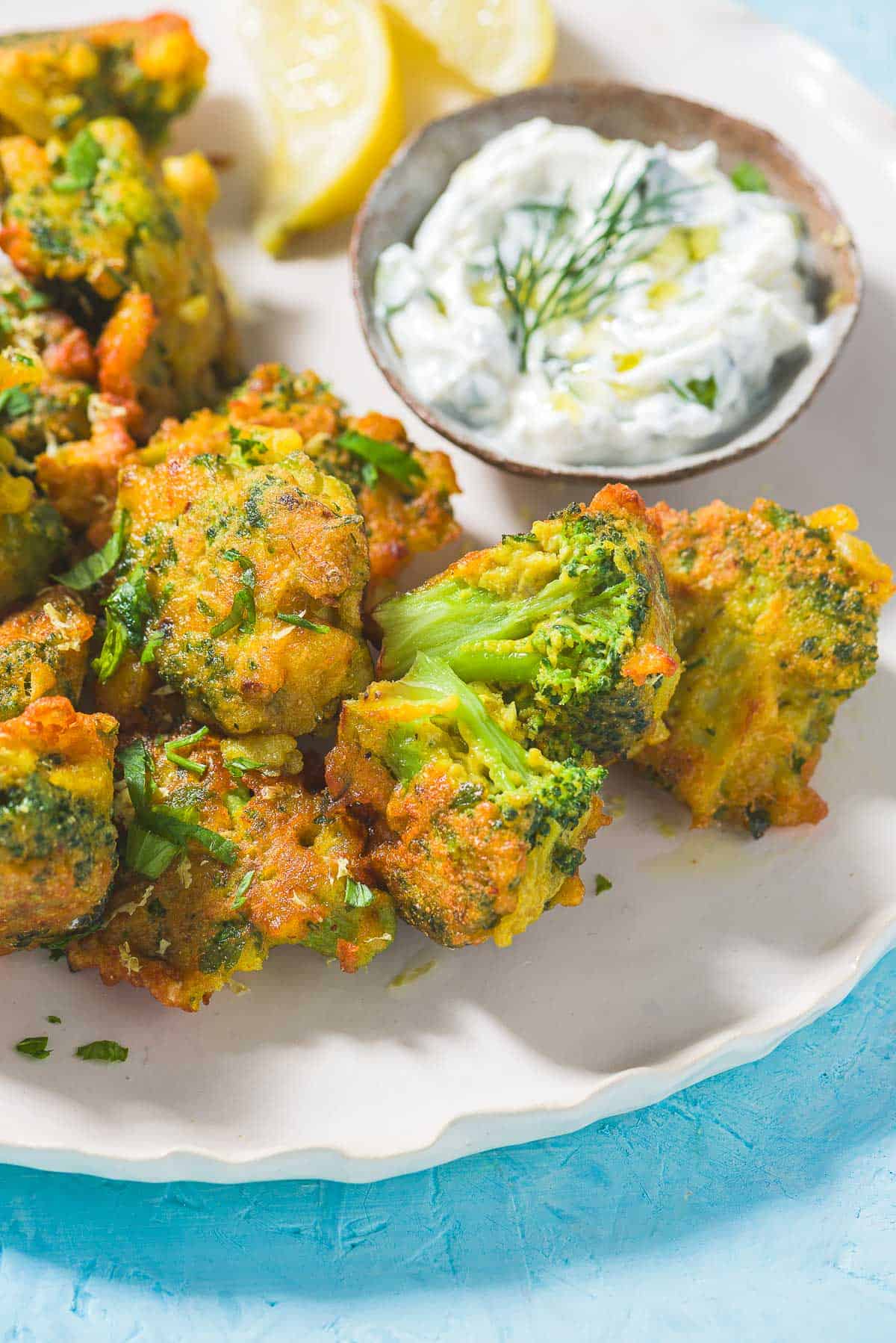 Broccoli Fritters on a plate with a small bowl of tzatziki sauce and lemon wedges.