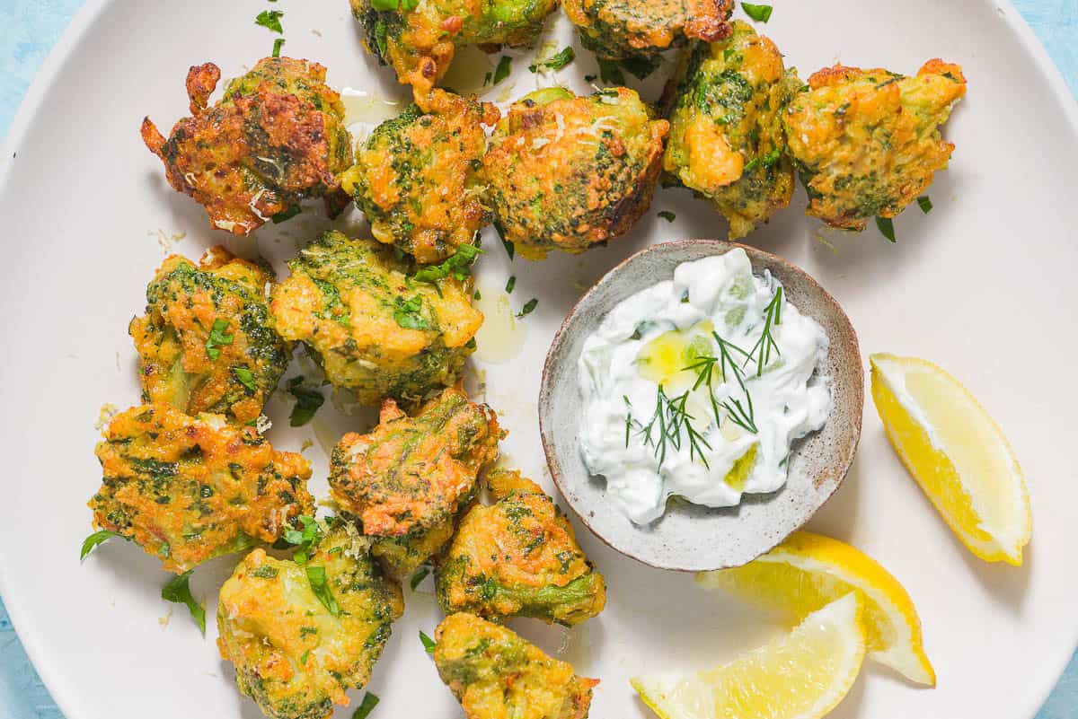 Broccoli Fritters on a plate with a small bowl of tzatziki sauce and lemon wedges.