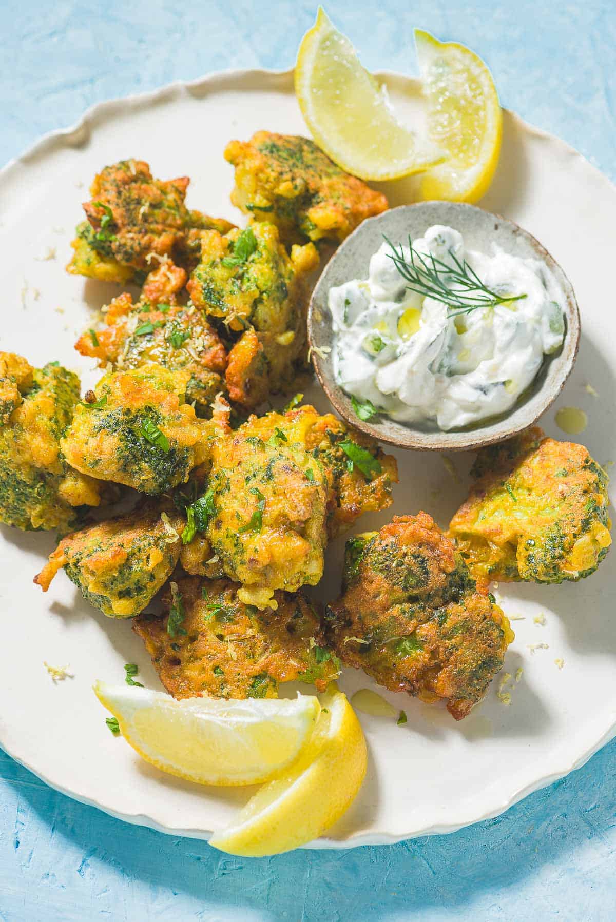Broccoli Fritters on a plate with a small bowl of tzatziki sauce and lemon wedges.