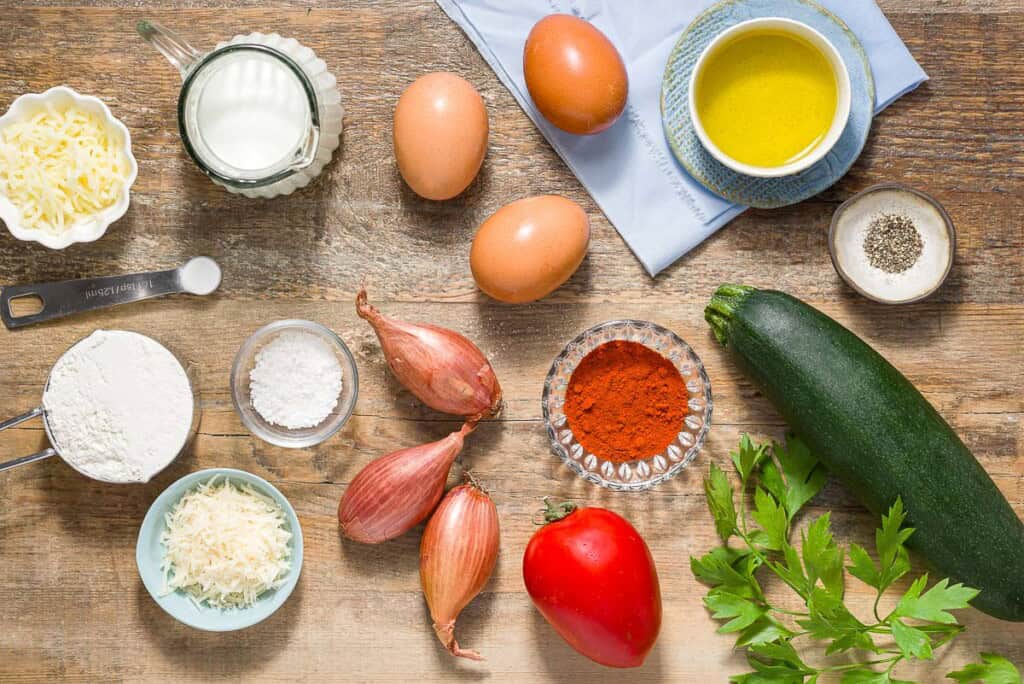 Ingredients for crustless zucchini quiche including eggs, tomato, olive oil, zucchini, shallots, sweet spanish paprika, salt, black pepper, shredded mozzarella, grated parmesan, milk, baking powder, white whole wheat flour and fresh parsley.