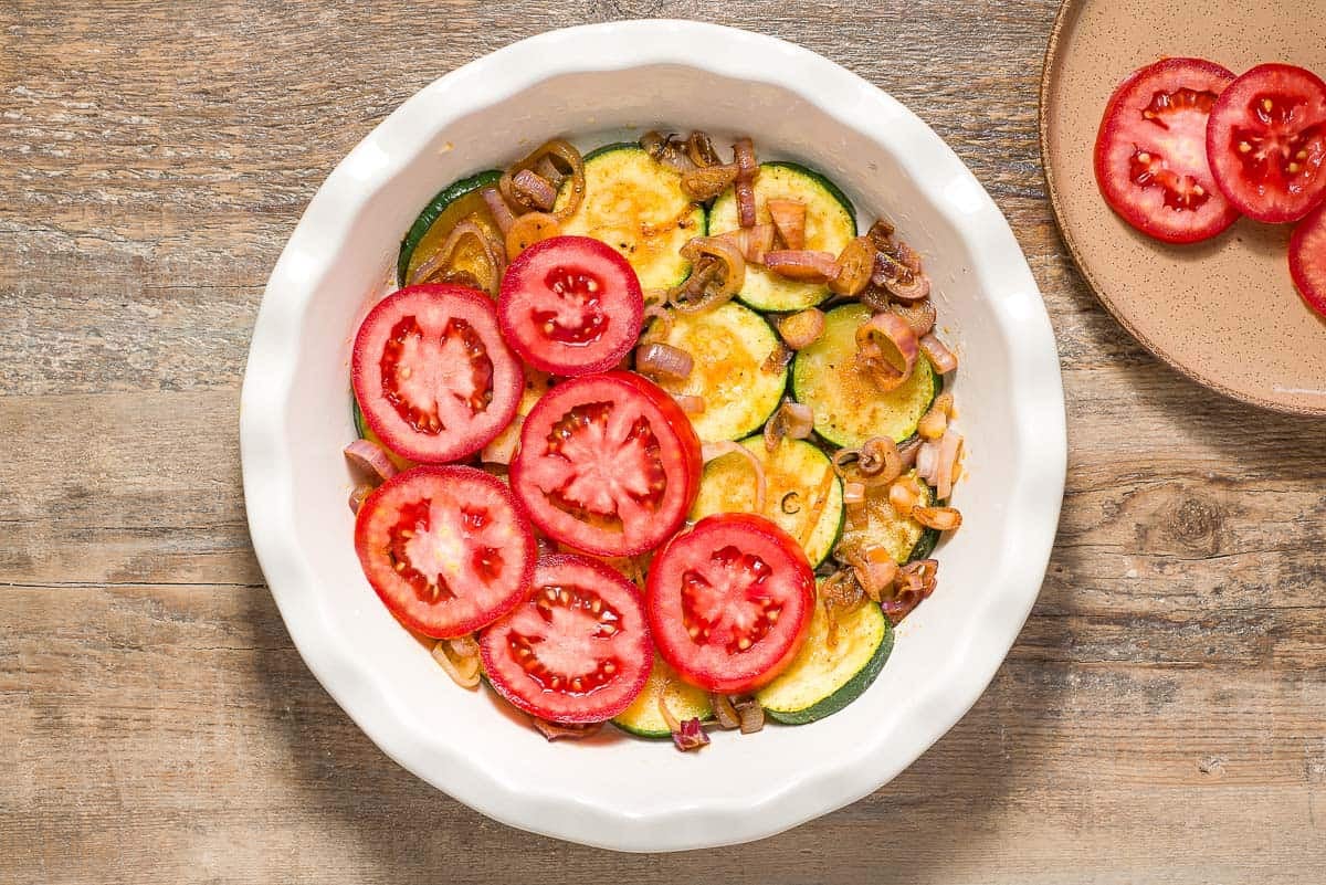 The vegetables for the crustless zucchini quiche in being layered in a baking dish. Next to this is a plate with some sliced tomatoes.