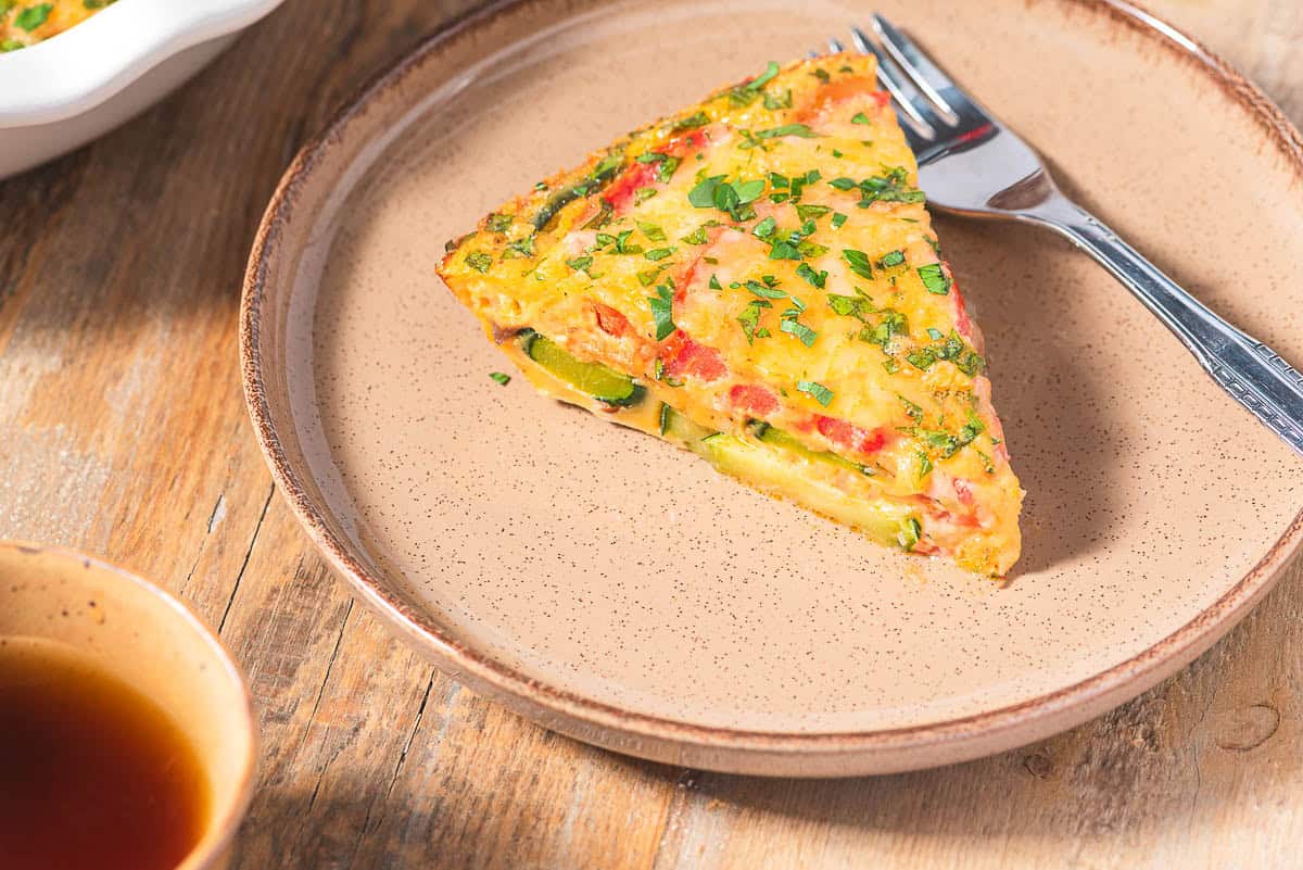 A slice of crustless zucchini quiche on a plate with a fork next to a cup of tea.