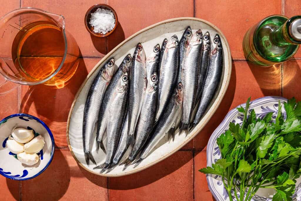 Ingredients for boquerones including fresh anchovies, white wine vinegar, olive oil, garlic, parsley and flaky sea salt.