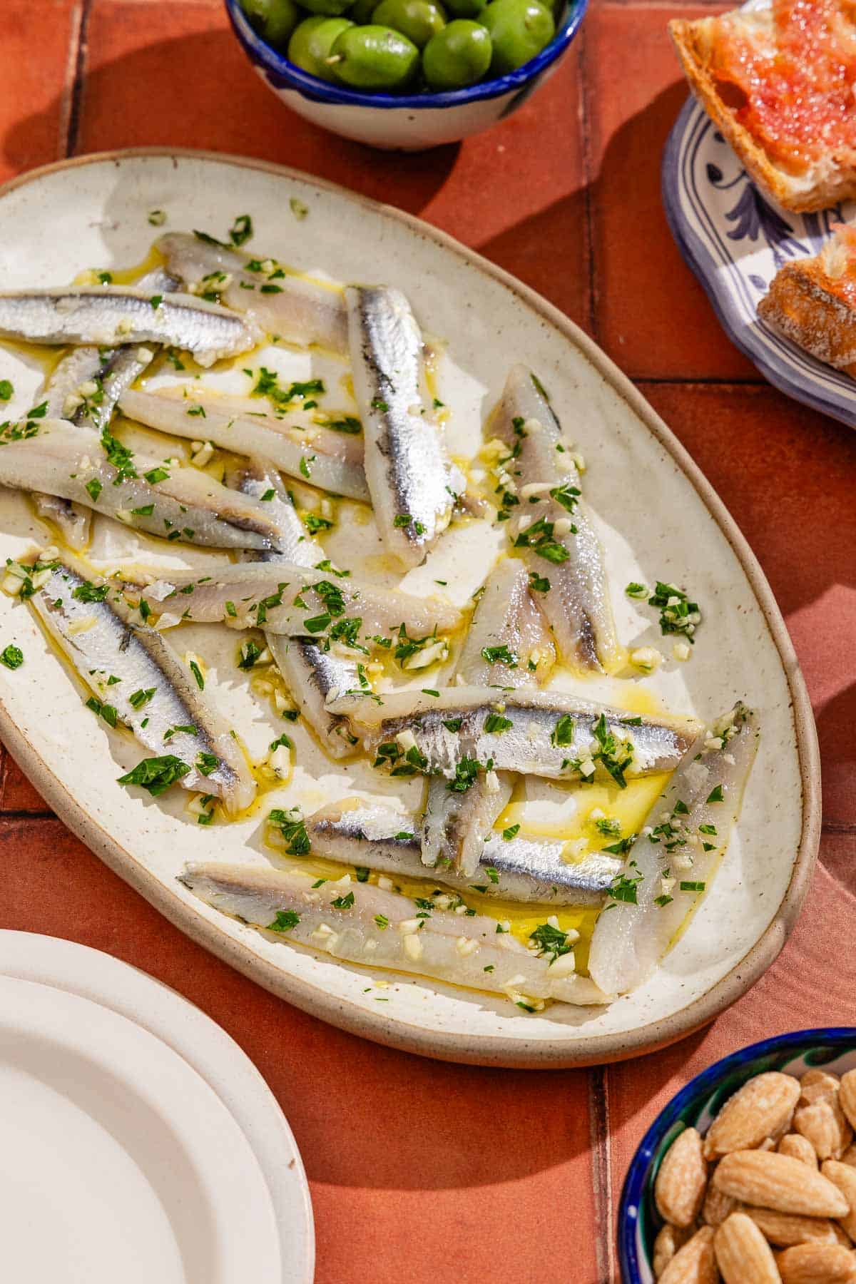 Boquerones on a platter next to a bowls of green olives and olives, a plate of pan con tomate, and a stack of 2 plates.