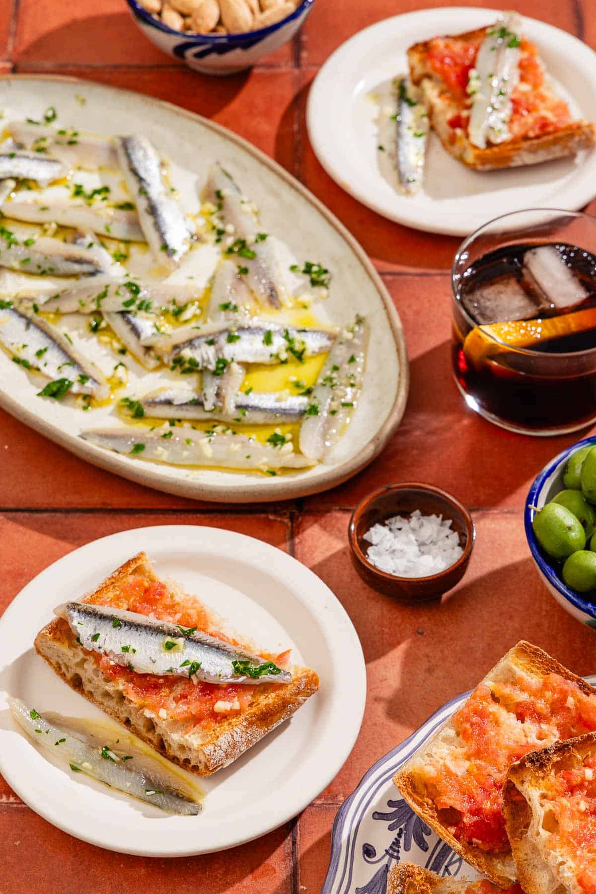 A platter of boquerones surrounded by two plates of boquerones on pan con tomate, bowls of flaky sea salt, almonds and green onions, a plate of pan con tomate and a glass of red sangria.