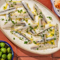 Boquerones on a platter next to a plate of pan con tomate, 2 bowls with green olives and almonds, and a stack of 2 plates.
