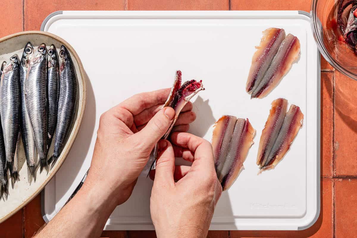 An anthovy being cleaned out over a cutting board with two anchovy fillets. Next to this is a platter of whole fresh anchovies.