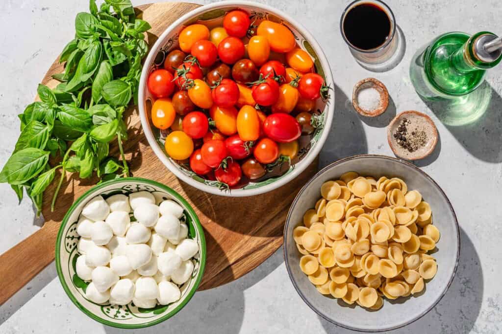 Ingredients for caprese pasta salad including ear pastry, salt, cherry tomatoes, mini mozzarella, olive oil, balsamic vinegar, black pepper and fresh basil.
